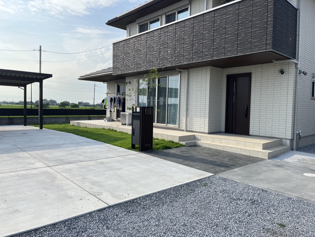 明るい色のレンガの外装と、2階に暗いアクセントのあるモダンな2階建ての家。前庭にはコンクリートと砂利の小道、小さな芝生、洗濯物が干してある物干しロープがあります。左側には屋根付きのカーポートエリアがあり、入り口に続く階段があります。