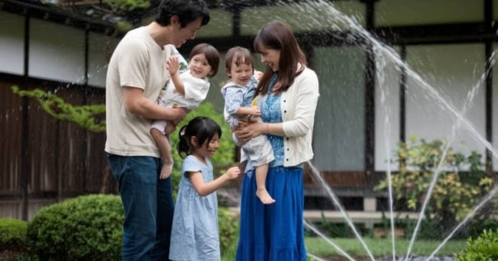 楽しそうな家族が、周囲に水が吹き出す庭の噴水の近くに立っています。ベージュのシャツを着た父親は、笑っている子供を抱いています。青いドレスを着た母親は、微笑みながらもう一人の子供を抱きかかえています。明るい色のドレスを着た三人目の子供は、彼らの間に立って、周囲の青々とした芝生に向かって手を伸ばしています。.