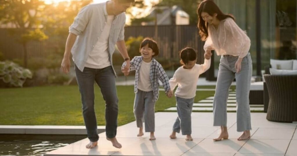 4 人家族がプールサイドで晴れた日を楽しんでいます。両親と幼い子供たちが手をつなぎ、手入れの行き届いた芝生の上を裸足で歩きながら笑顔を見せています。背景の緑と金色の太陽が、暖かく楽しい雰囲気を作り出しています。.