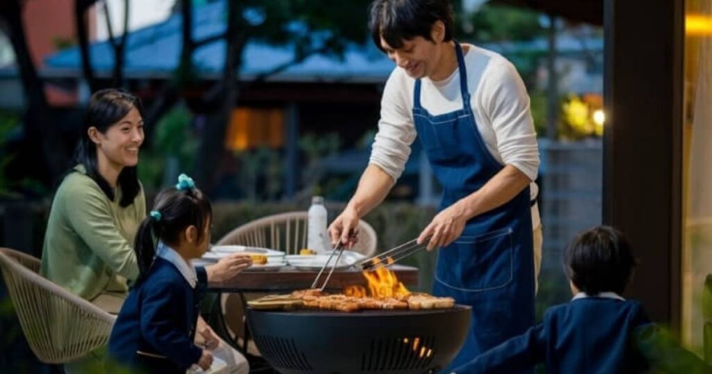 エプロンをつけた男性が屋外の丸いバーベキューグリルで肉を焼いている。近くのテーブルには皿とボトルが置かれたが、そこに女性と2人の子供が笑顔で座っている。手入れの行き届いた庭の芝生は薄暗い光の中で輝き、暖かく居心地の良い雰囲気を醸し出している。.