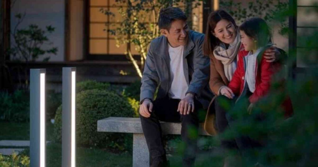 3 人家族が、美しく整えられた庭の石のベンチに夜座っています。灰色のジャケットを着た父親と、茶色のコートとスカーフを身に着けた母親が、赤いジャケットを着た娘に温かく微笑んでいます。2 つの垂直のライトが、茂みと家を背景にした初心者向けのこのシーンを照らしています。.