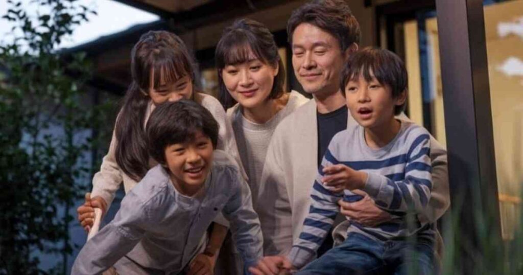 5人家族がテラスの外に座り、笑顔を見せています。両親の両脇には男の子2人と女の子1人の3人の子供がいます。子供たちはカジュアルな服装で、背景には木々と建物があり、日中の家族の温かいひとときをとらえています。.